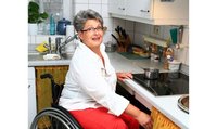  Une femme aux cheveux courts gris et portant des lunettes est assise dans un fauteuil roulant devant la cuisinière dans sa cuisine.