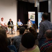 Die ersten paar Reihen des Publikums von hinten, eine Person steht und hält ein Mikrofon in der Hand. Fünf Personen auf der Bühne blicken die Person im Publikum an.