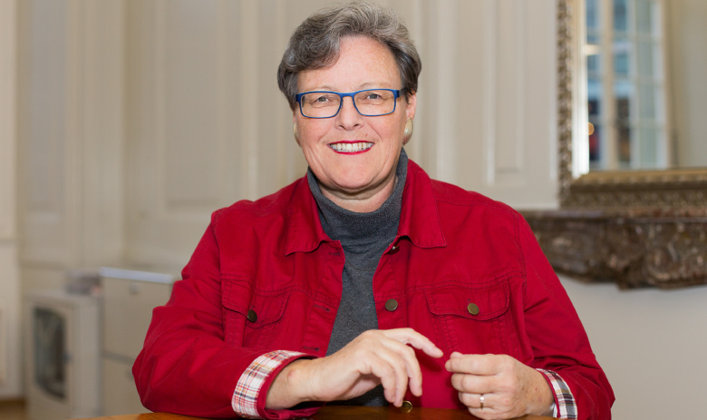 Annekäthi B. Portraitfoto einer Frau, die lächelnd in die Kamera blickt. Sie trägt eine rote Jacke, darunter einen grauen Rollkragenpullover.