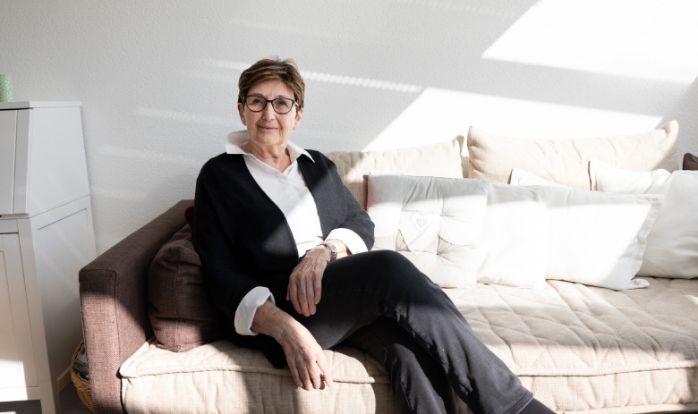 Josiane P Eine Frau sitzt auf einem Sofa. Das Zimmer ist sehr hell eingerichtet, sie selbst trägt dunkle Kleidung.