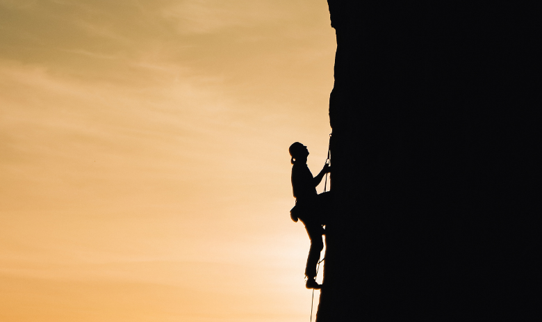 Silhouette einer Person, die eine steile Felswand heraufklettert, vor einem Sonnenaufgang.